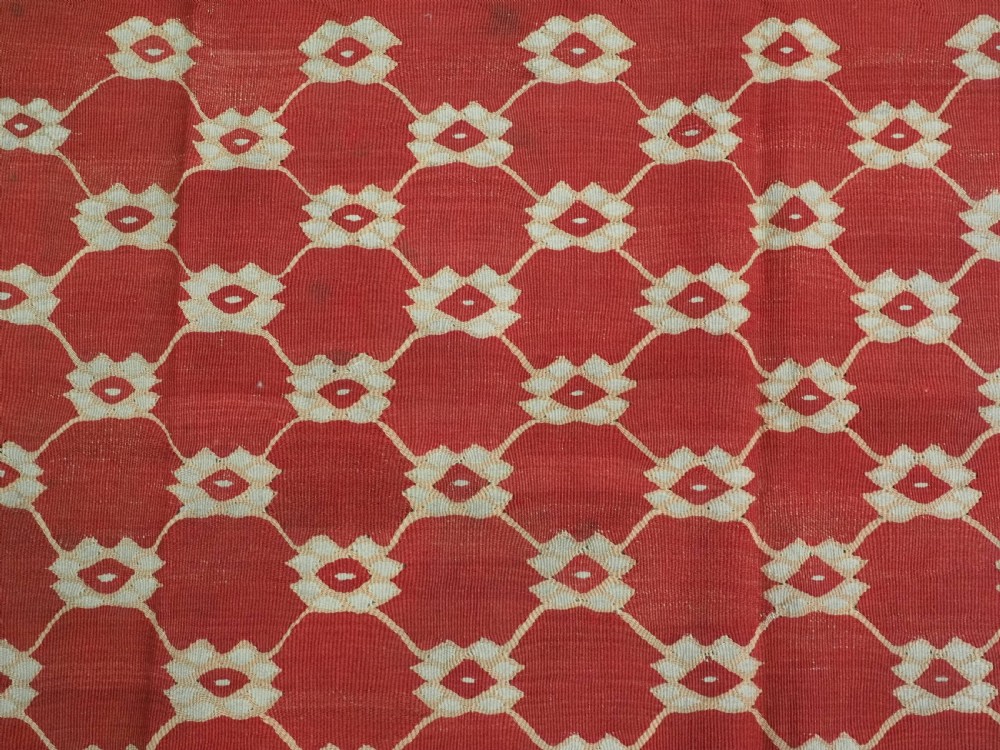 A rectangular flatweave wool kilim carpet featuring a red centre field with a cream lattice pattern, enclosed by a geometric cream and red border. View VIII