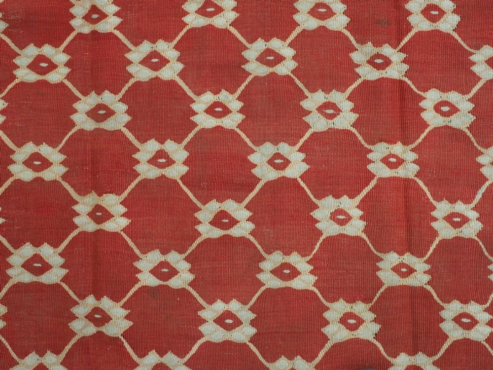 A rectangular flatweave wool kilim carpet featuring a red centre field with a cream lattice pattern, enclosed by a geometric cream and red border. View IX