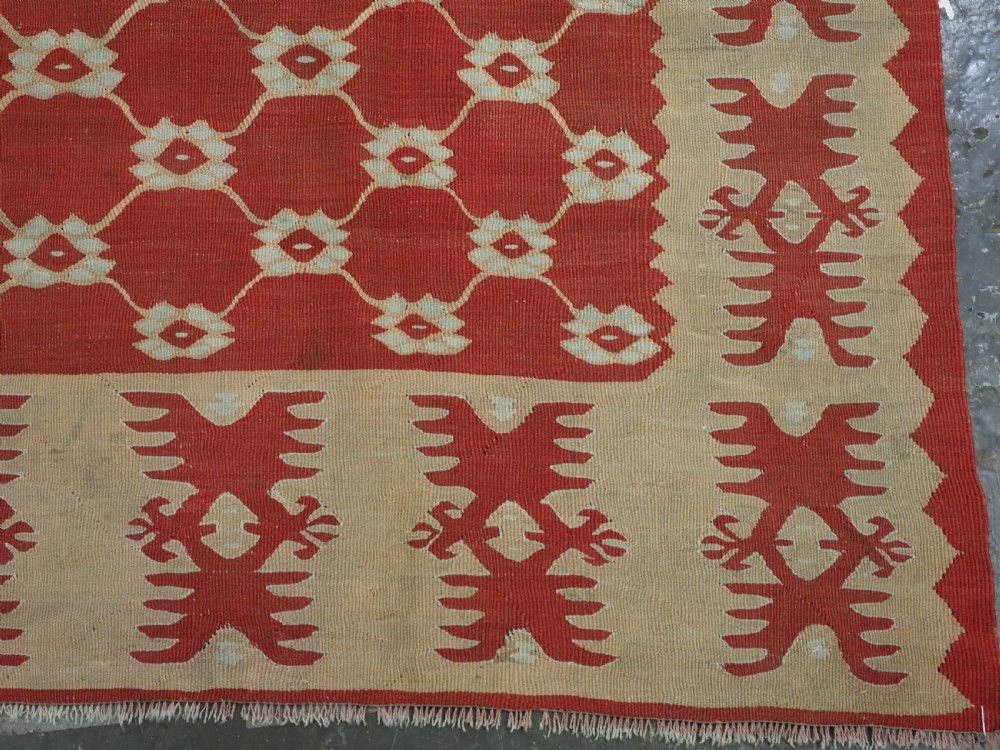 A rectangular flatweave wool kilim carpet featuring a red centre field with a cream lattice pattern, enclosed by a geometric cream and red border. View X