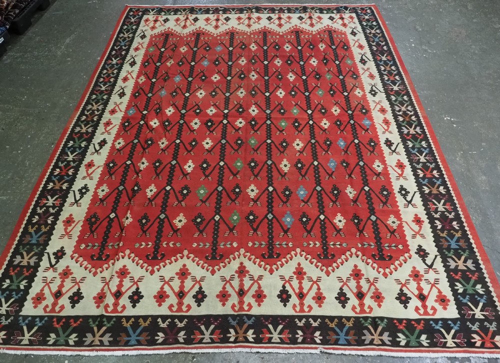 An antique flatweave wool kilim carpet featuring a central red field patterned with vertical flowering tree motifs surrounded by layered geometric borders.