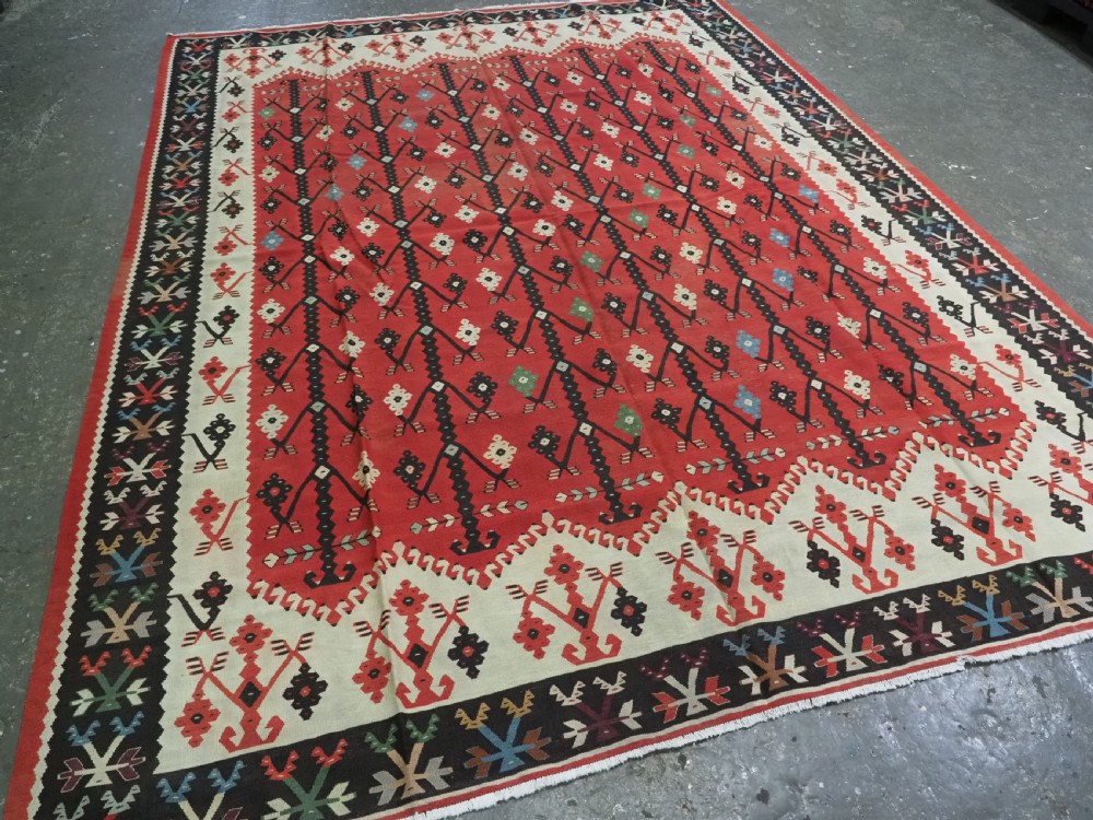 An antique flatweave wool kilim carpet featuring a central red field patterned with vertical flowering tree motifs surrounded by layered geometric borders. View II