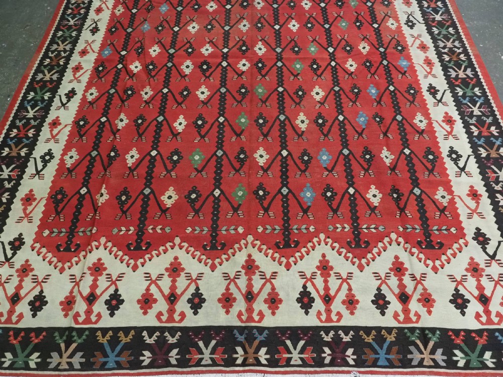 An antique flatweave wool kilim carpet featuring a central red field patterned with vertical flowering tree motifs surrounded by layered geometric borders. View V