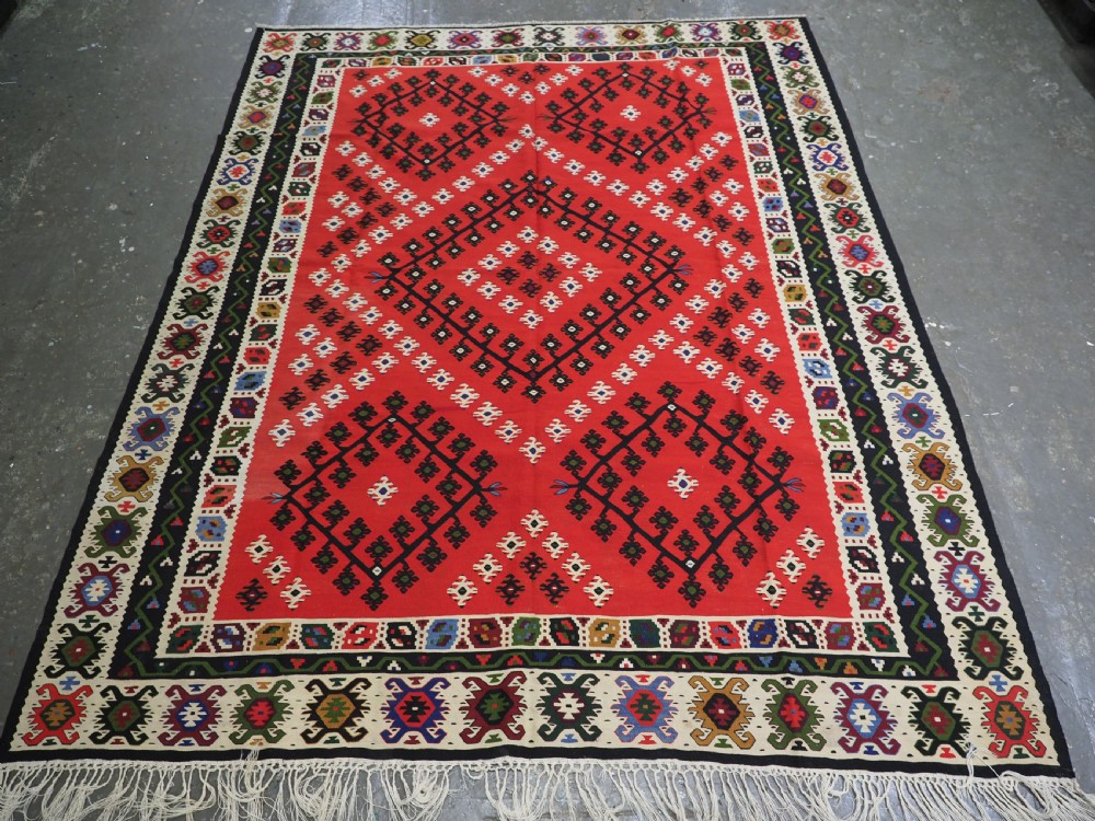 An antique red Pirot Sarkoy wool kilim rug featuring black and white geometric diamond motifs and multi-coloured decorative borders.