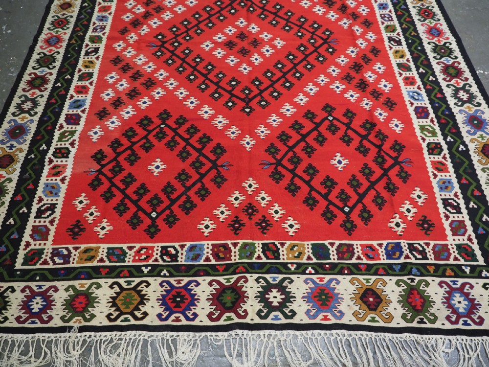 An antique red Pirot Sarkoy wool kilim rug featuring black and white geometric diamond motifs and multi-coloured decorative borders. View V
