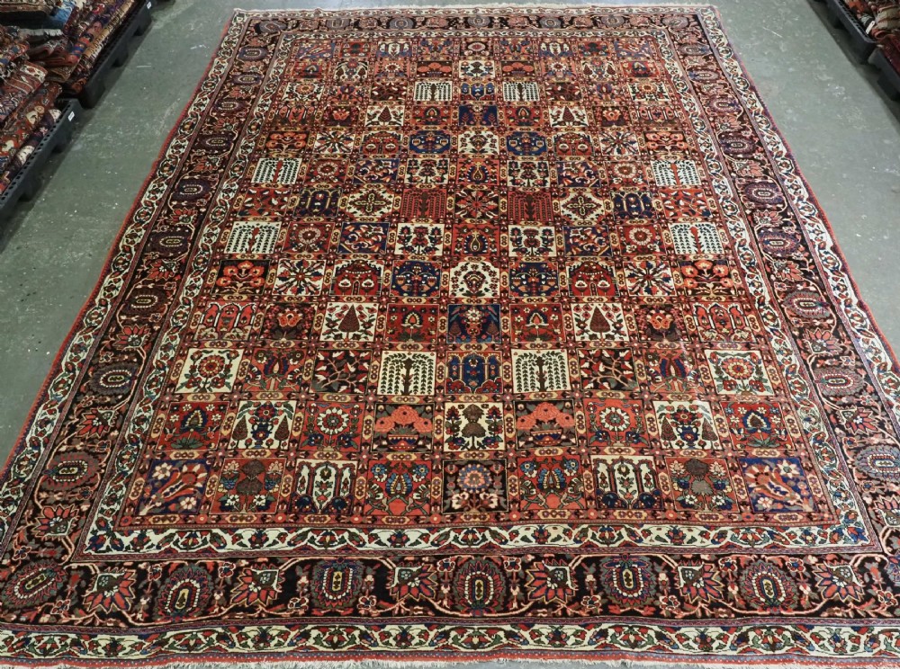 A large antique Bakhtiari wool carpet featuring a repeating grid pattern of garden motifs in terracotta red, navy blue, and ivory framed by a wide ornamental border.