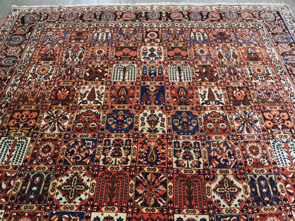 A large antique Bakhtiari wool carpet featuring a repeating grid pattern of garden motifs in terracotta red, navy blue, and ivory framed by a wide ornamental border. View III