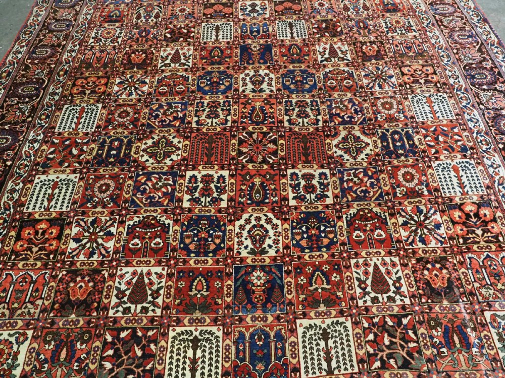A large antique Bakhtiari wool carpet featuring a repeating grid pattern of garden motifs in terracotta red, navy blue, and ivory framed by a wide ornamental border. View IV