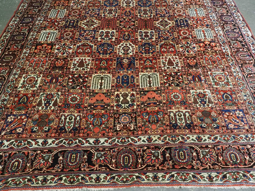 A large antique Bakhtiari wool carpet featuring a repeating grid pattern of garden motifs in terracotta red, navy blue, and ivory framed by a wide ornamental border. View V