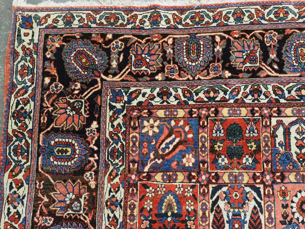 A large antique Bakhtiari wool carpet featuring a repeating grid pattern of garden motifs in terracotta red, navy blue, and ivory framed by a wide ornamental border. View VI
