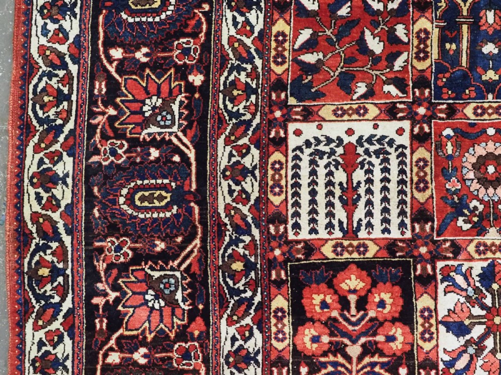A large antique Bakhtiari wool carpet featuring a repeating grid pattern of garden motifs in terracotta red, navy blue, and ivory framed by a wide ornamental border. View VII