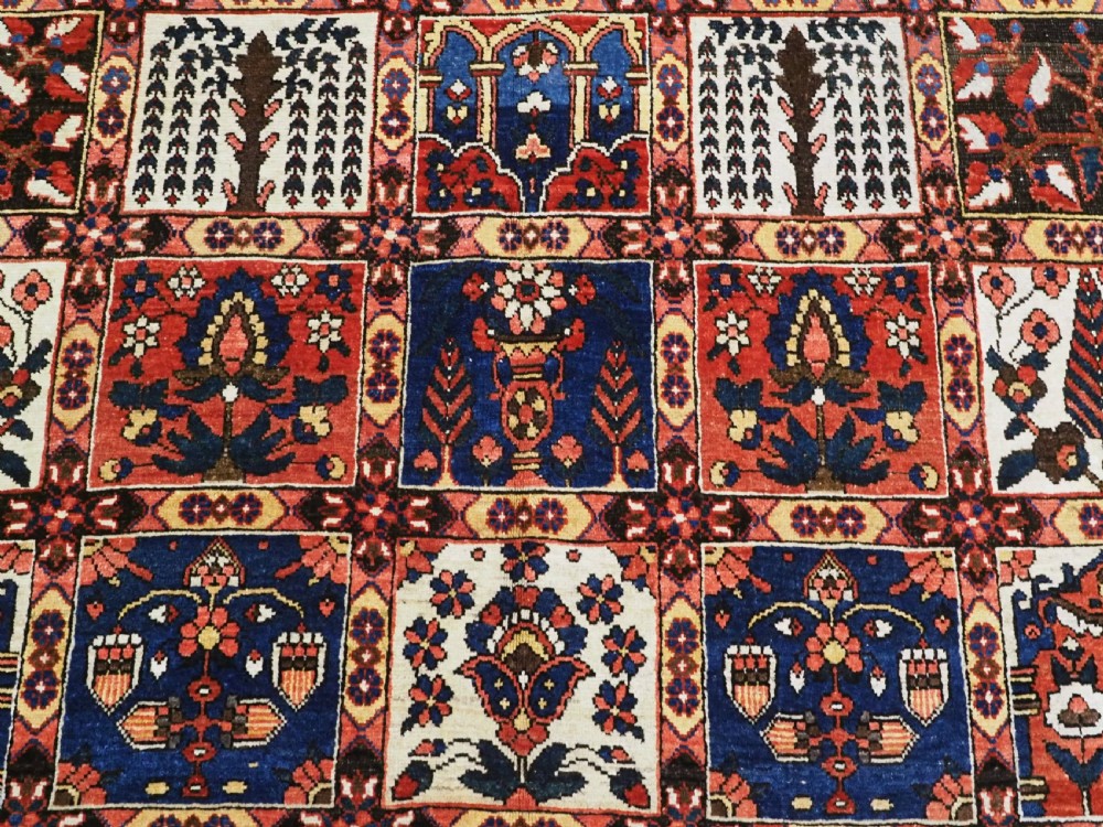 A large antique Bakhtiari wool carpet featuring a repeating grid pattern of garden motifs in terracotta red, navy blue, and ivory framed by a wide ornamental border. View IX