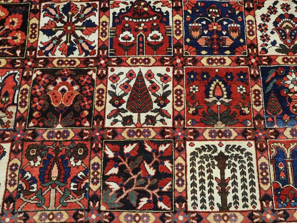 A large antique Bakhtiari wool carpet featuring a repeating grid pattern of garden motifs in terracotta red, navy blue, and ivory framed by a wide ornamental border. View X
