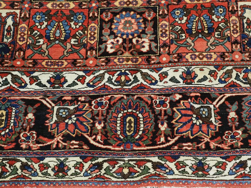 A large antique Bakhtiari wool carpet featuring a repeating grid pattern of garden motifs in terracotta red, navy blue, and ivory framed by a wide ornamental border. View XII
