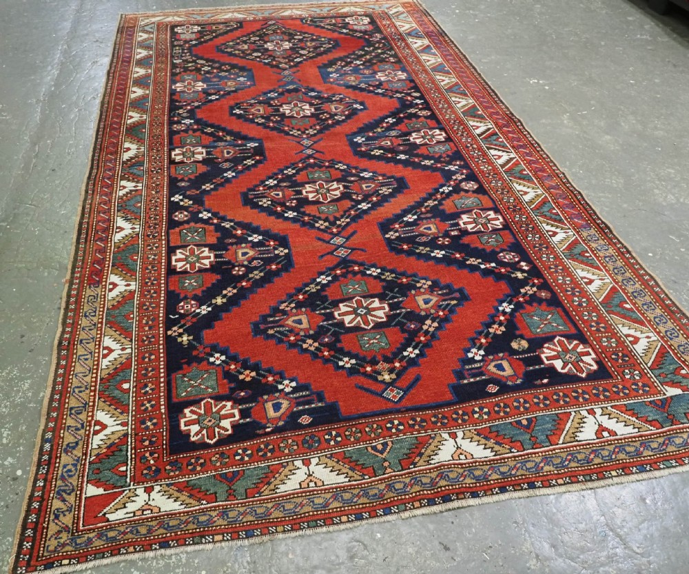 An antique Caucasian Kazak kelleh rug with a central red field, blue geometric diamond medallions, and multi-layered patterned borders. View II