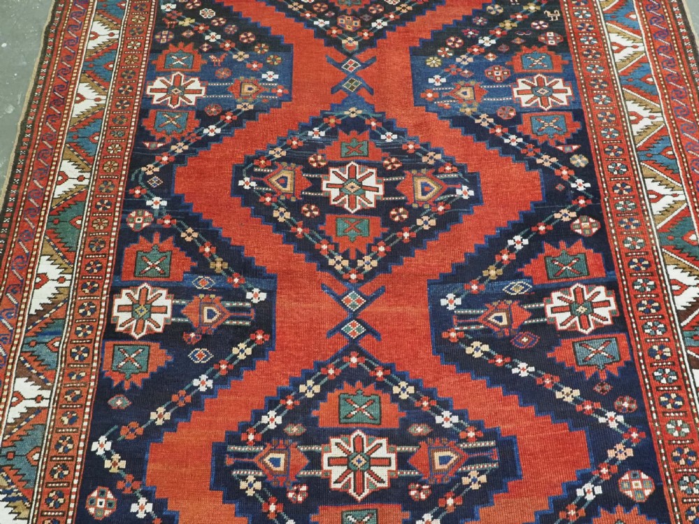 An antique Caucasian Kazak kelleh rug with a central red field, blue geometric diamond medallions, and multi-layered patterned borders. View IV