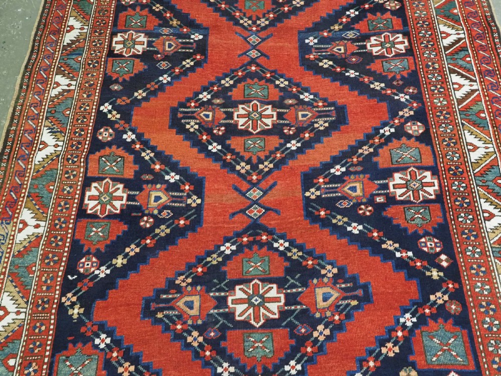 An antique Caucasian Kazak kelleh rug with a central red field, blue geometric diamond medallions, and multi-layered patterned borders. View V