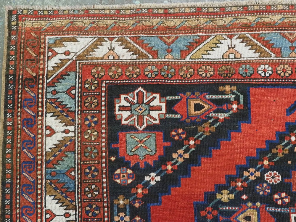An antique Caucasian Kazak kelleh rug with a central red field, blue geometric diamond medallions, and multi-layered patterned borders. View VI