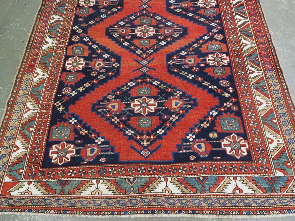 An antique Caucasian Kazak kelleh rug with a central red field, blue geometric diamond medallions, and multi-layered patterned borders. View VII