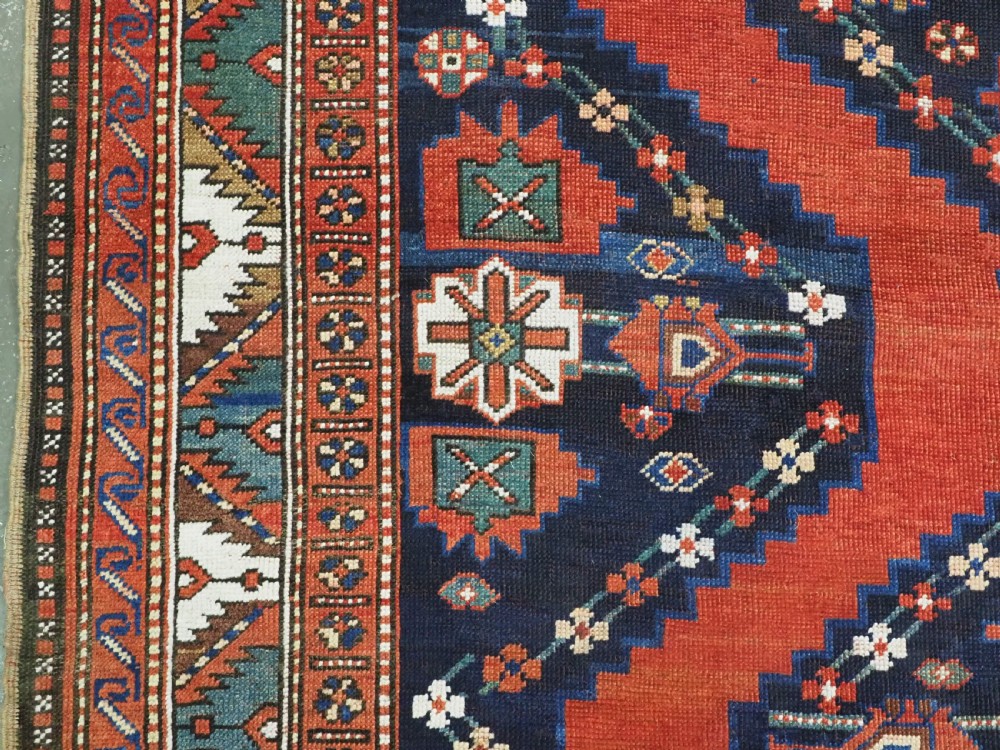 An antique Caucasian Kazak kelleh rug with a central red field, blue geometric diamond medallions, and multi-layered patterned borders. View VIII