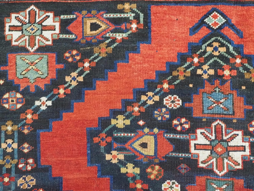 An antique Caucasian Kazak kelleh rug with a central red field, blue geometric diamond medallions, and multi-layered patterned borders. View IX