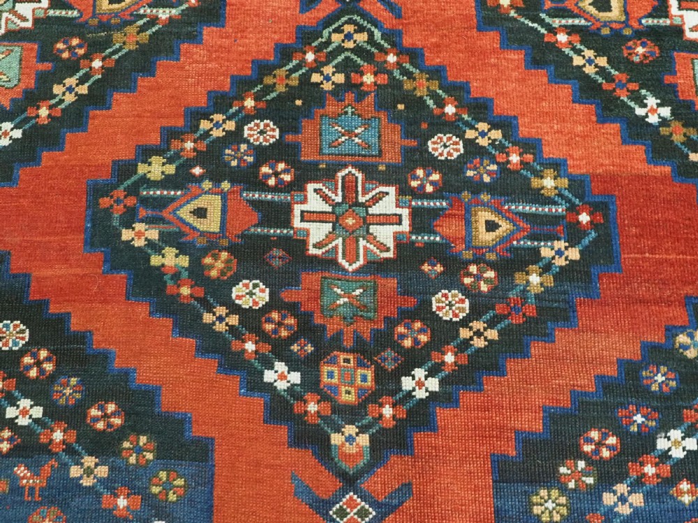 An antique Caucasian Kazak kelleh rug with a central red field, blue geometric diamond medallions, and multi-layered patterned borders. View X