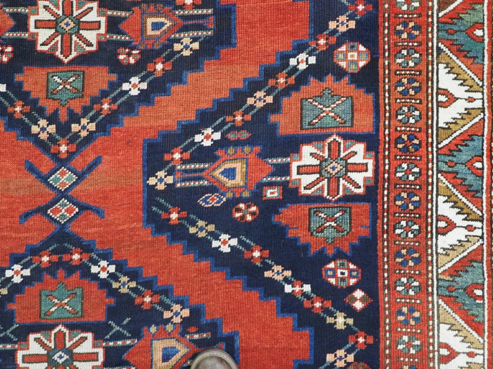 An antique Caucasian Kazak kelleh rug with a central red field, blue geometric diamond medallions, and multi-layered patterned borders. View XIII