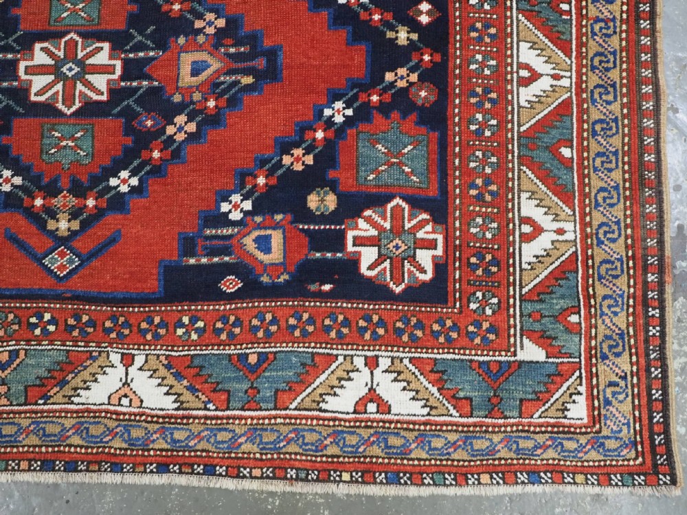An antique Caucasian Kazak kelleh rug with a central red field, blue geometric diamond medallions, and multi-layered patterned borders. View XIV