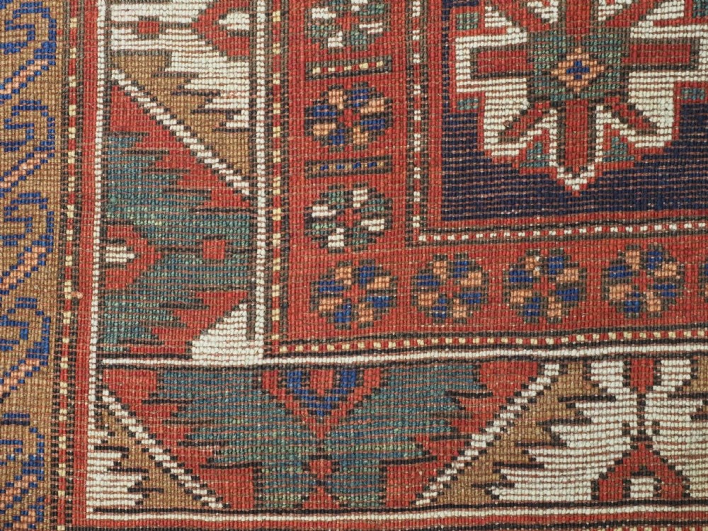 An antique Caucasian Kazak kelleh rug with a central red field, blue geometric diamond medallions, and multi-layered patterned borders. View XV