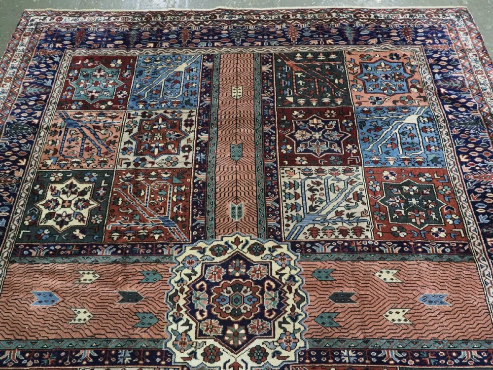 A rectangular hand-knotted wool carpet featuring a central grid of garden panels framed by a wide dark navy geometric border in terracotta, blue, and cream tones. View III