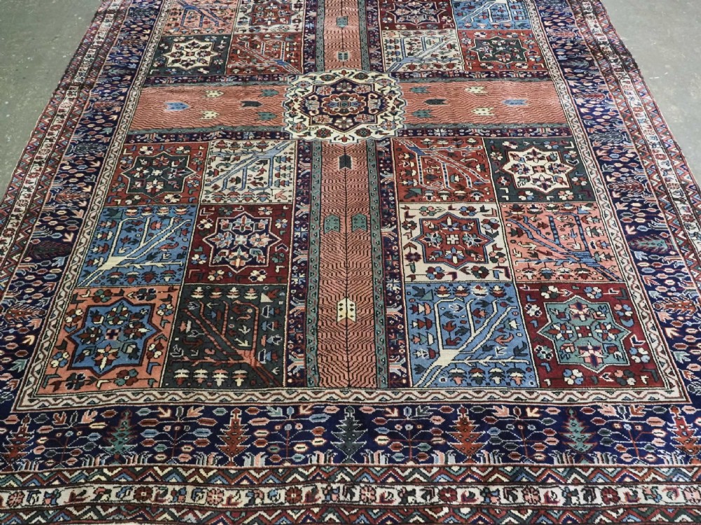 A rectangular hand-knotted wool carpet featuring a central grid of garden panels framed by a wide dark navy geometric border in terracotta, blue, and cream tones. View V