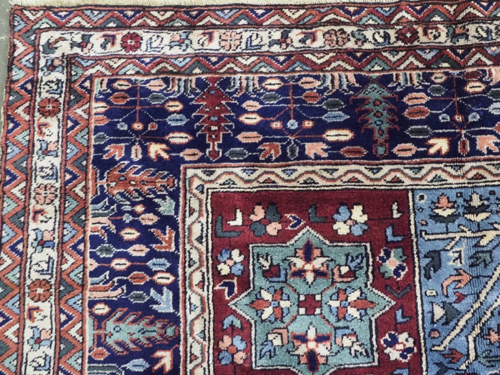 A rectangular hand-knotted wool carpet featuring a central grid of garden panels framed by a wide dark navy geometric border in terracotta, blue, and cream tones. View VI