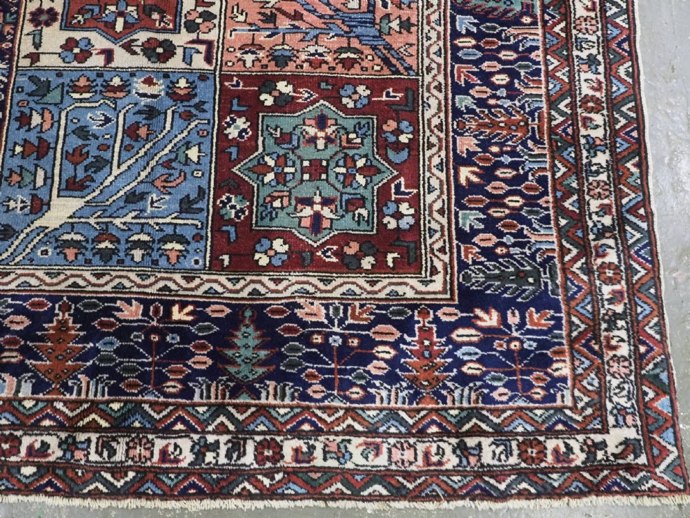 A rectangular hand-knotted wool carpet featuring a central grid of garden panels framed by a wide dark navy geometric border in terracotta, blue, and cream tones. View XII