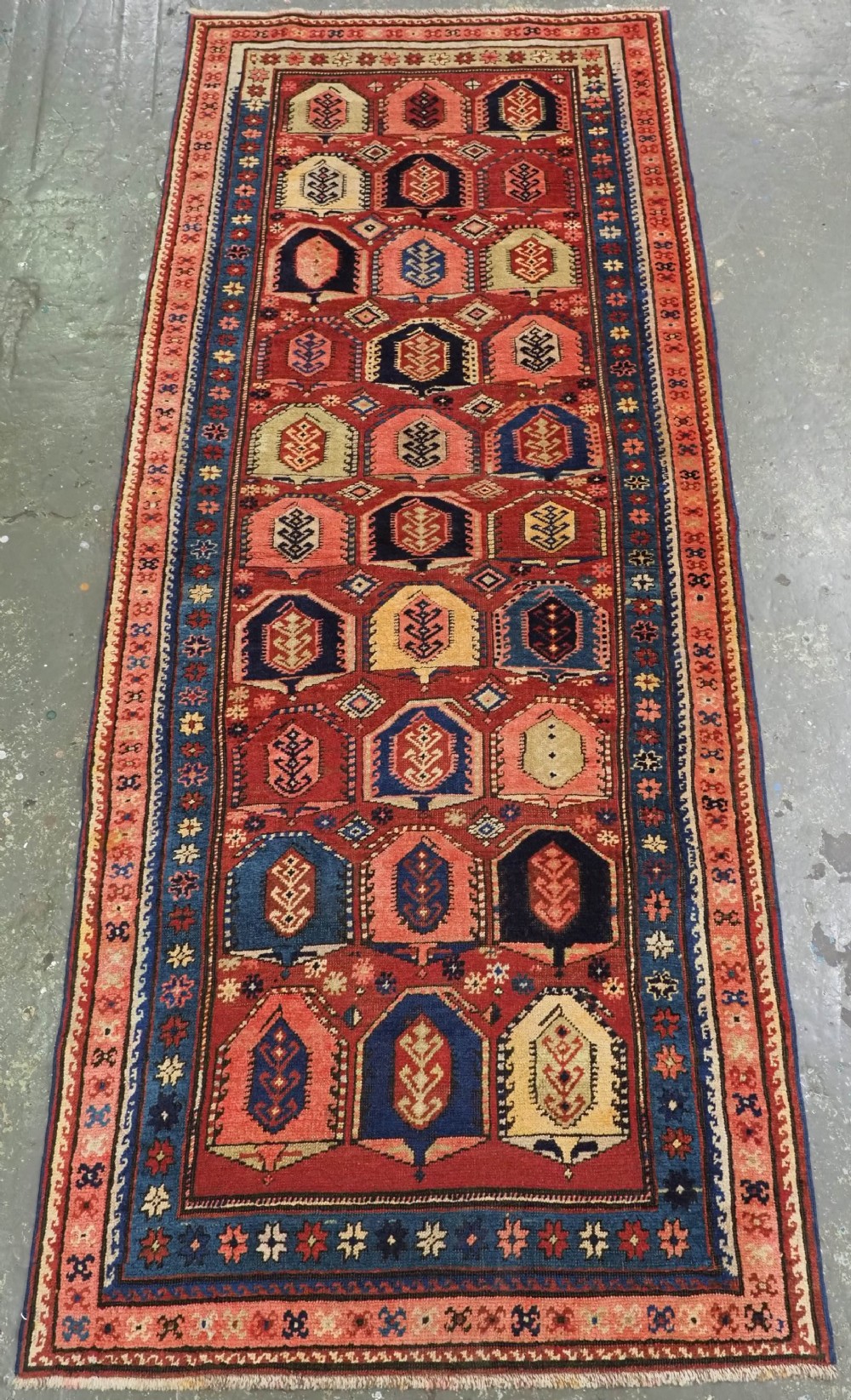 An antique Caucasian long rug featuring repeating geometric house-shaped boteh motifs in blue, cream, and pink across a terracotta red field with decorative multi-band borders.