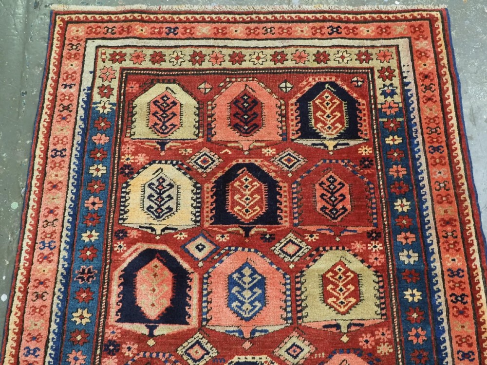 An antique Caucasian long rug featuring repeating geometric house-shaped boteh motifs in blue, cream, and pink across a terracotta red field with decorative multi-band borders. View III