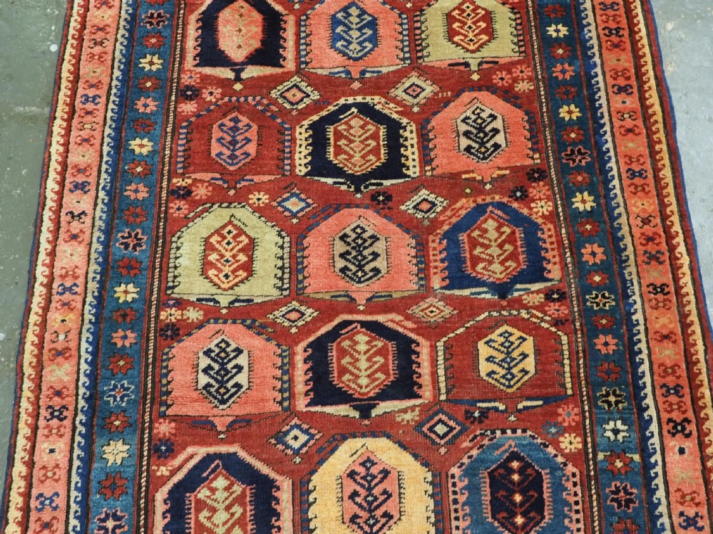 An antique Caucasian long rug featuring repeating geometric house-shaped boteh motifs in blue, cream, and pink across a terracotta red field with decorative multi-band borders. View IV