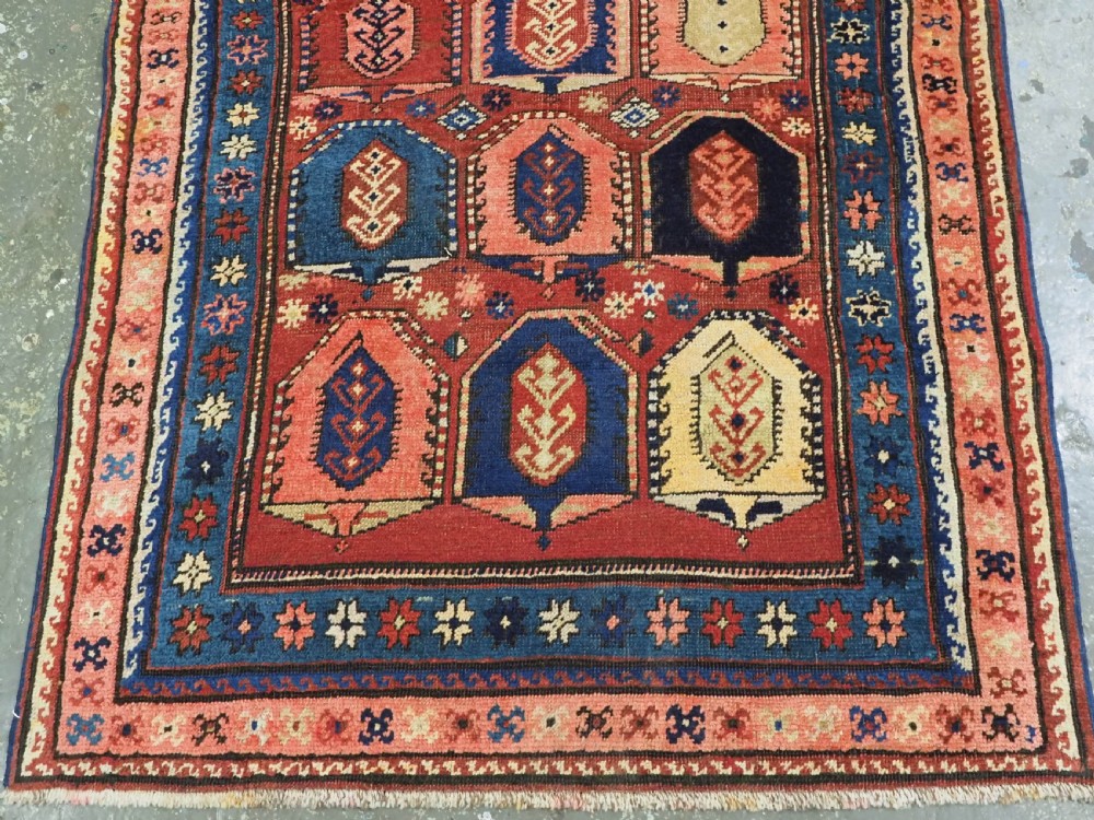 An antique Caucasian long rug featuring repeating geometric house-shaped boteh motifs in blue, cream, and pink across a terracotta red field with decorative multi-band borders. View V