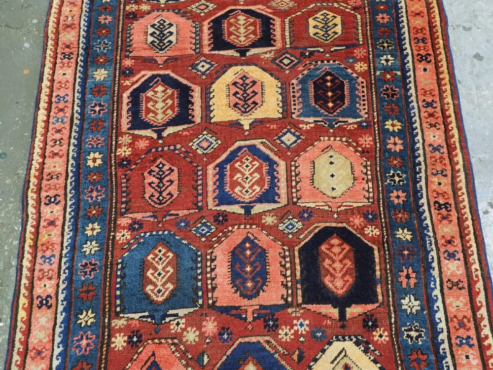 An antique Caucasian long rug featuring repeating geometric house-shaped boteh motifs in blue, cream, and pink across a terracotta red field with decorative multi-band borders. View VI