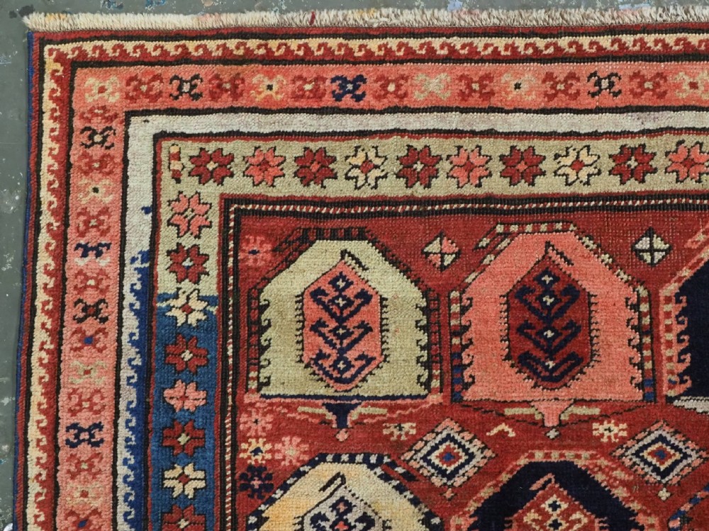 An antique Caucasian long rug featuring repeating geometric house-shaped boteh motifs in blue, cream, and pink across a terracotta red field with decorative multi-band borders. View VII