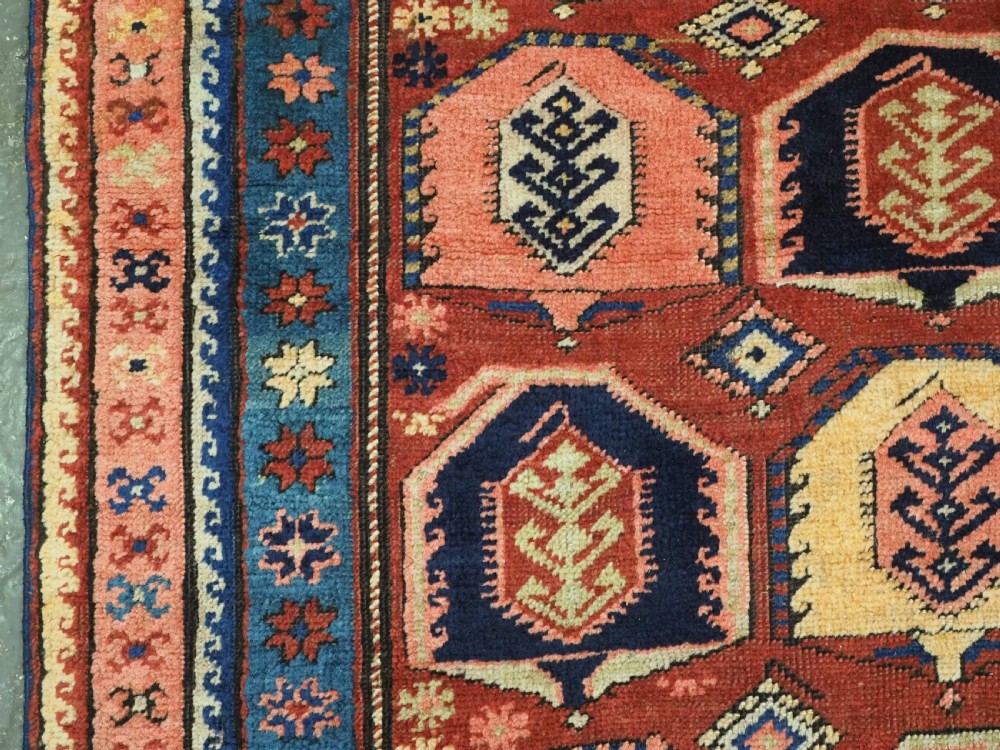 An antique Caucasian long rug featuring repeating geometric house-shaped boteh motifs in blue, cream, and pink across a terracotta red field with decorative multi-band borders. View VIII