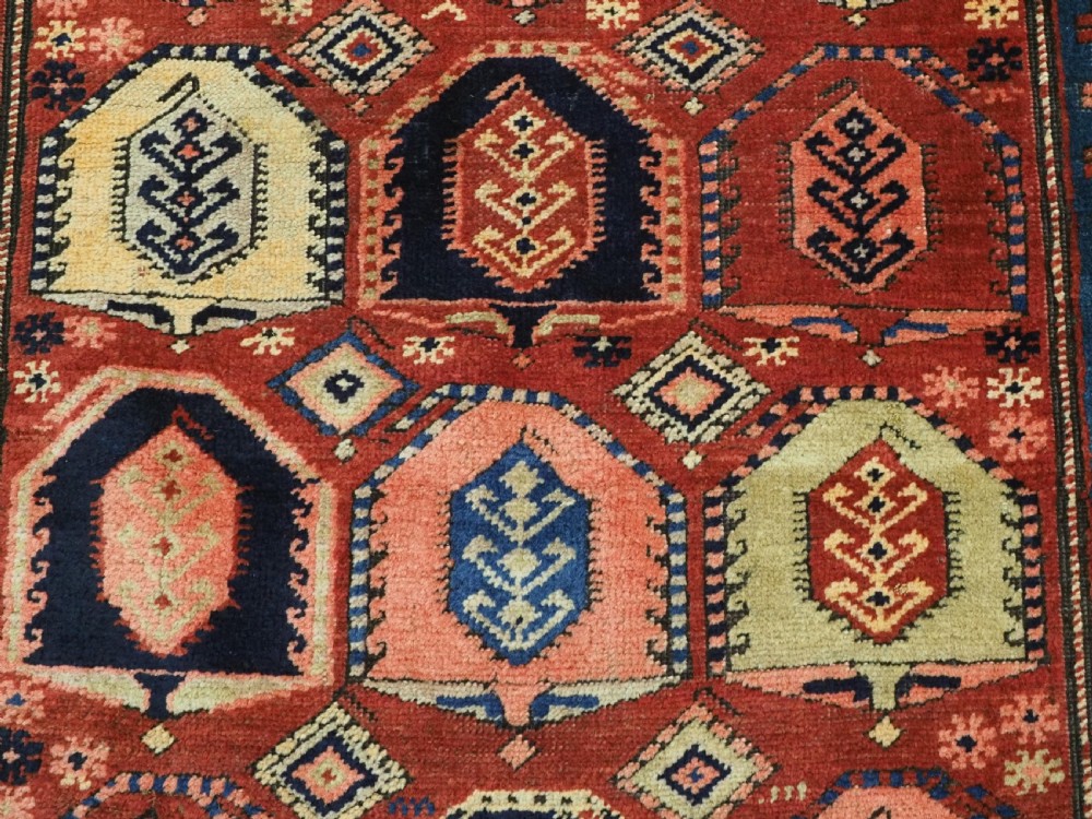 An antique Caucasian long rug featuring repeating geometric house-shaped boteh motifs in blue, cream, and pink across a terracotta red field with decorative multi-band borders. View IX