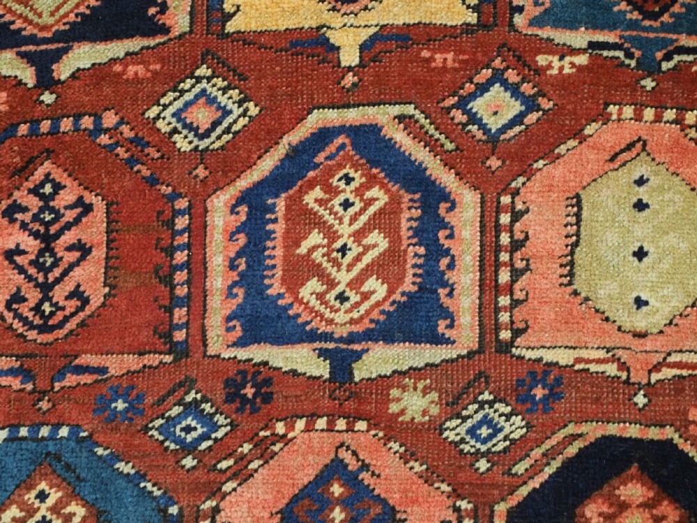 An antique Caucasian long rug featuring repeating geometric house-shaped boteh motifs in blue, cream, and pink across a terracotta red field with decorative multi-band borders. View X