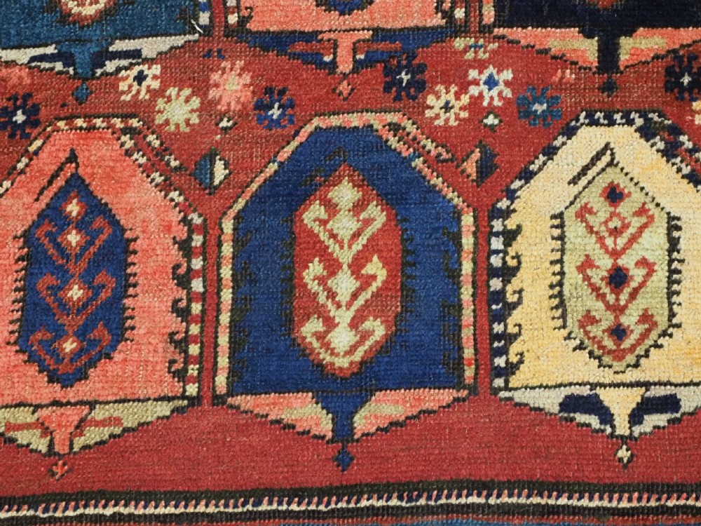 An antique Caucasian long rug featuring repeating geometric house-shaped boteh motifs in blue, cream, and pink across a terracotta red field with decorative multi-band borders. View XI