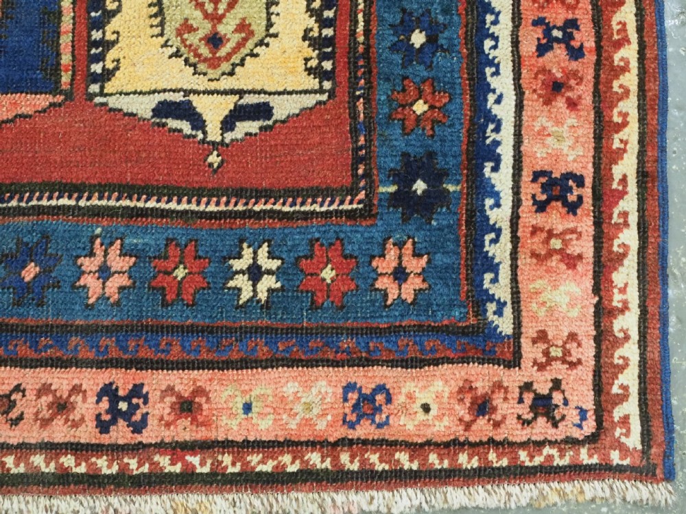 An antique Caucasian long rug featuring repeating geometric house-shaped boteh motifs in blue, cream, and pink across a terracotta red field with decorative multi-band borders. View XII