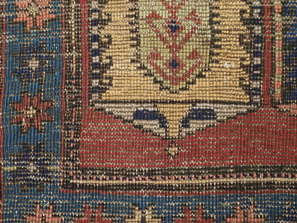 An antique Caucasian long rug featuring repeating geometric house-shaped boteh motifs in blue, cream, and pink across a terracotta red field with decorative multi-band borders. View XIII
