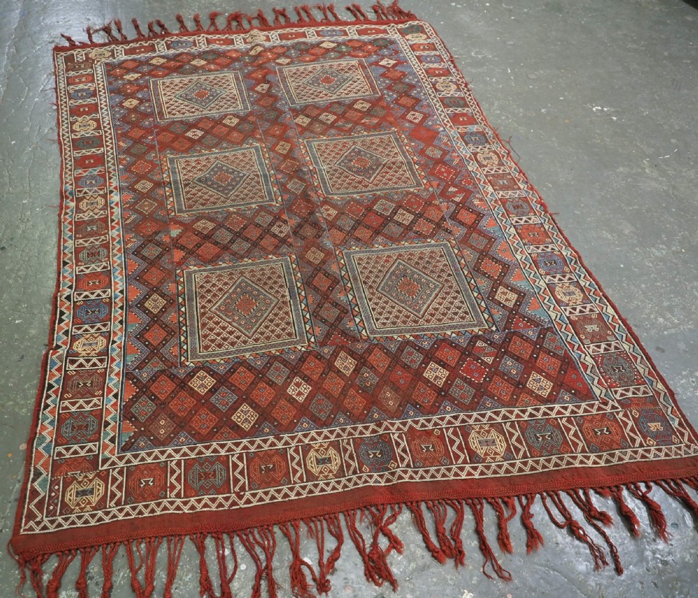 An antique Caucasian flatweave rug displaying geometric diamond medallions in rust red, navy blue, and cream wool with fringed ends. View II