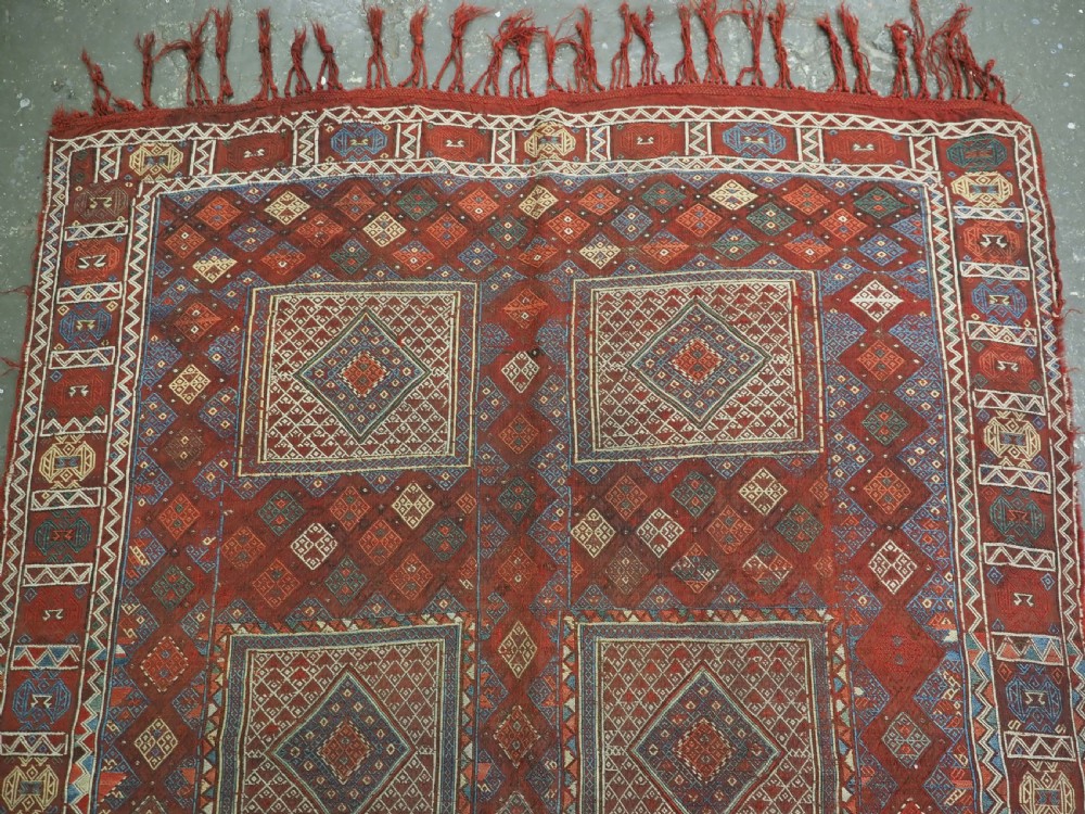 An antique Caucasian flatweave rug displaying geometric diamond medallions in rust red, navy blue, and cream wool with fringed ends. View III