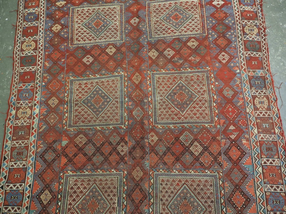An antique Caucasian flatweave rug displaying geometric diamond medallions in rust red, navy blue, and cream wool with fringed ends. View IV