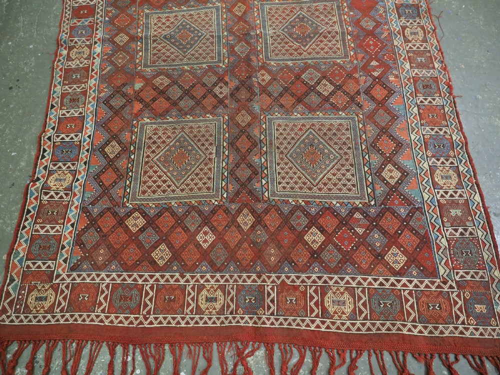 An antique Caucasian flatweave rug displaying geometric diamond medallions in rust red, navy blue, and cream wool with fringed ends. View V