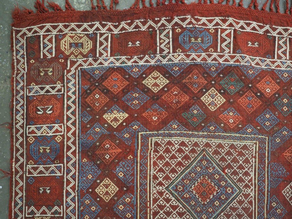 An antique Caucasian flatweave rug displaying geometric diamond medallions in rust red, navy blue, and cream wool with fringed ends. View VI