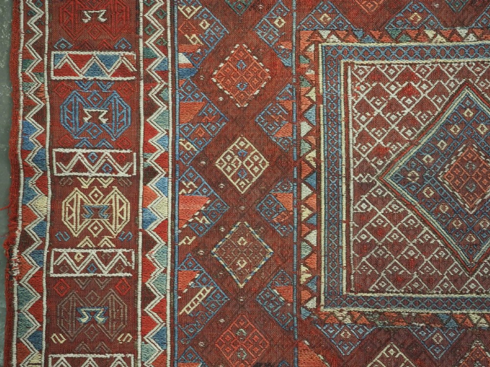 An antique Caucasian flatweave rug displaying geometric diamond medallions in rust red, navy blue, and cream wool with fringed ends. View VII
